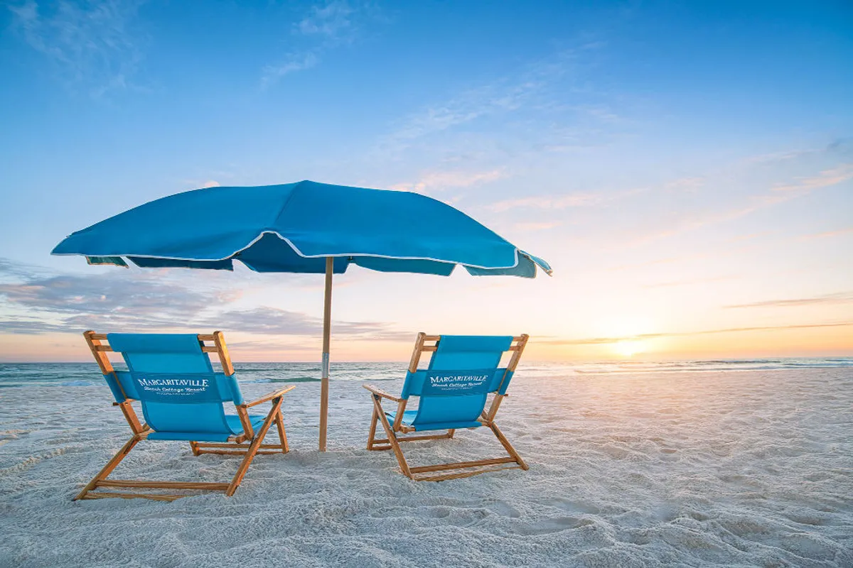 The image showcases two inviting blue beach chairs with 'Margaritaville Beach Cottage Resort' branding under a large blue umbrella, set on pristine white sand facing a breathtaking sunset over the Gulf of Mexico. The scene embodies relaxation, with the gentle waves and golden sky creating a tranquil atmosphere perfect for unwinding after a day of beachside activities.