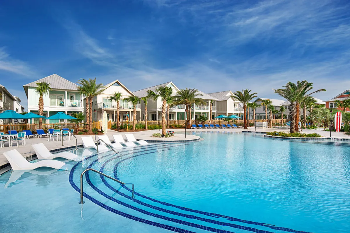 This wide-angle shot showcases the large heated oasis pool surrounded by modern beachfront cottages with balconies. Lush palm trees and tropical landscaping frame the scene, with guests swimming, lounging under blue umbrellas, and socializing at shaded tables.