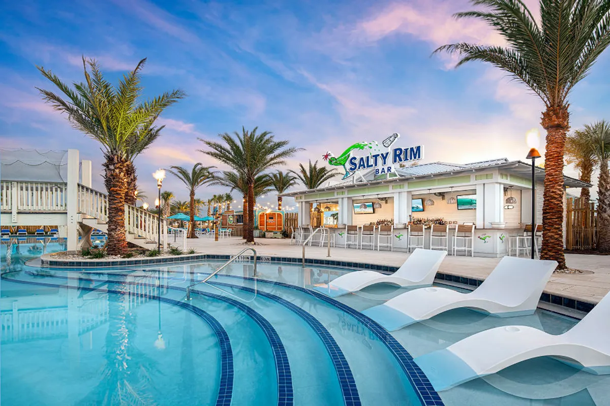 The image features the lively Salty Rim Bar with open seating adjacent to a curved pool with submerged lounge chairs. Palm trees surround the area, creating a festive, tropical atmosphere. Guests are seen swimming, lounging, and enjoying drinks and food at the poolside bar. A water slide and an elevated deck add to the sense of fun and activity.