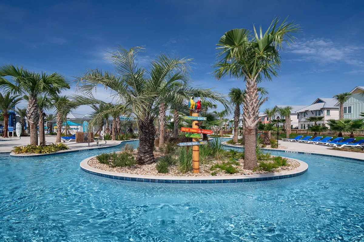 This image showcases a vibrant lazy river winding through lush tropical landscaping, dotted with palm trees and bordered by blue lounge chairs. A colorful signpost featuring playful tropical bird decorations adds a whimsical touch, while children and families can be seen floating and playing in the water, creating a joyful, carefree atmosphere.