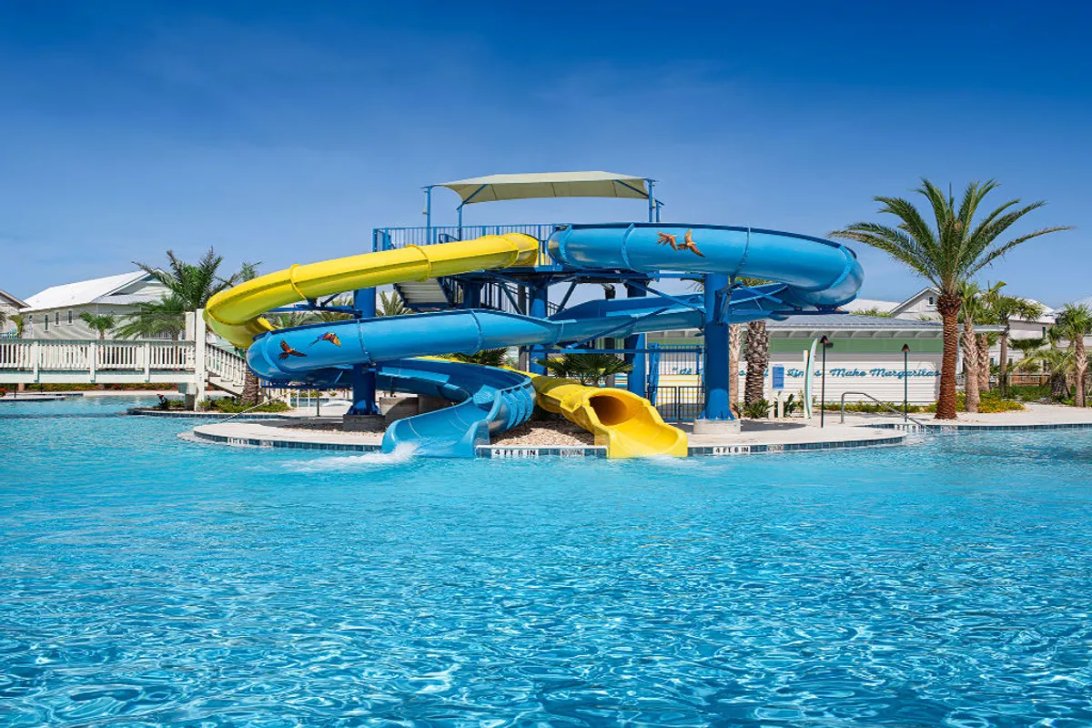 A dynamic pool scene featuring multi-level yellow and blue waterslides that curve into a large, sparkling swimming pool. The area is framed by tropical palm trees and shaded canopies, with families and friends enjoying the water, lounging, and gathering together.