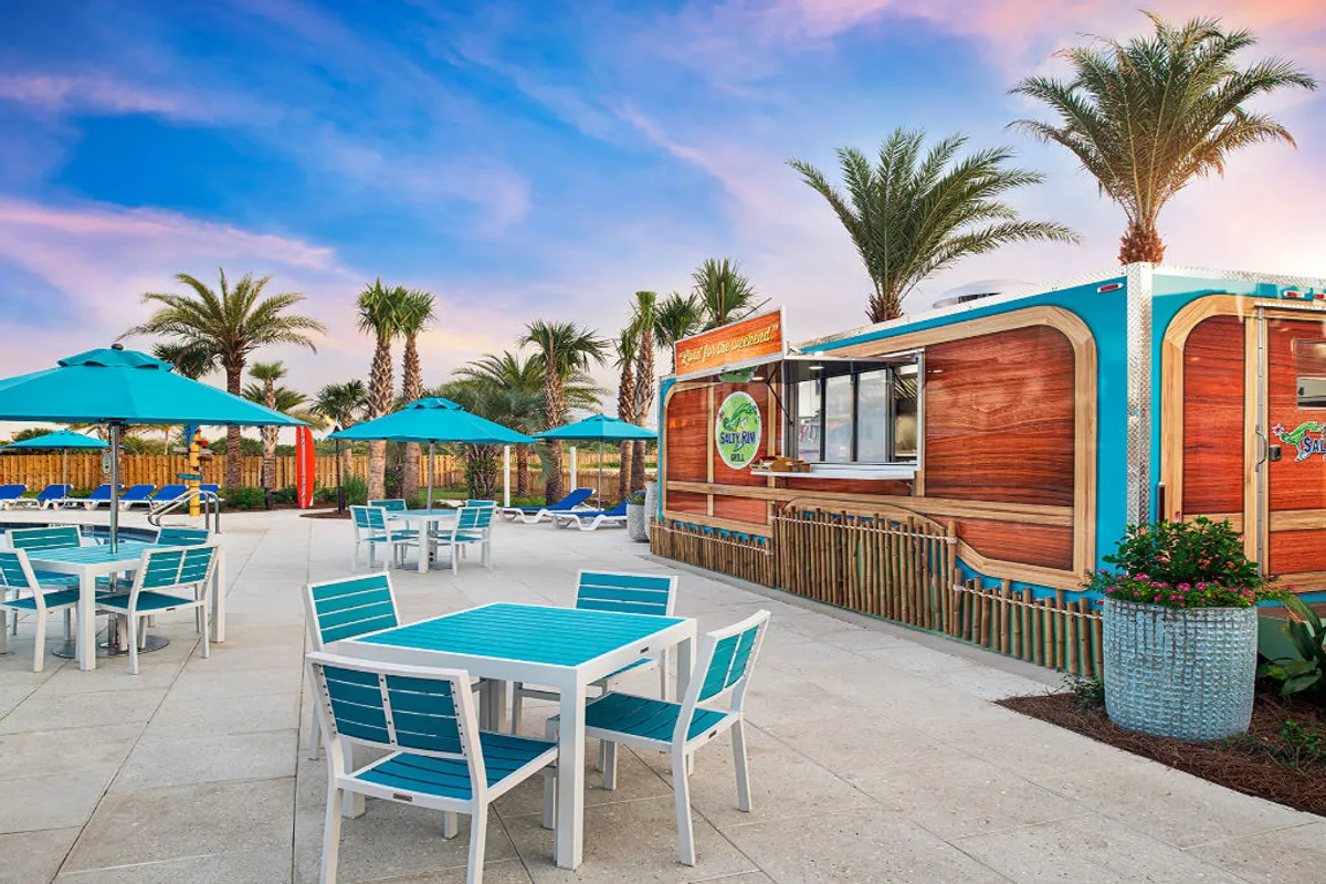 The image presents turquoise outdoor tables with matching umbrellas set beside a wood-paneled poolside bar marked by colorful signage. Tall palm trees and sun loungers complete the lively scene, where guests dine, relax, and socialize in a cheerful, carefree setting.