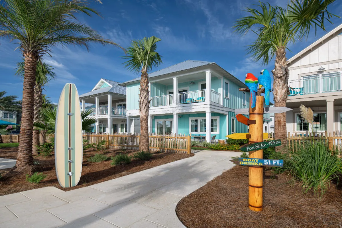 A vibrant exterior shot of a turquoise beach cottage, complete with inviting balconies adorned with surfboard accents and surrounded by lush tropical palm trees. In the foreground, a whimsical parrot signpost sets a cheerful tone, while guests can be seen relaxing on balconies and strolling along paved pathways that weave through the landscaped resort grounds. The cottage is just steps from the white sand beach and sparkling Gulf waters, with hints of beach and water activities visible in the distance.