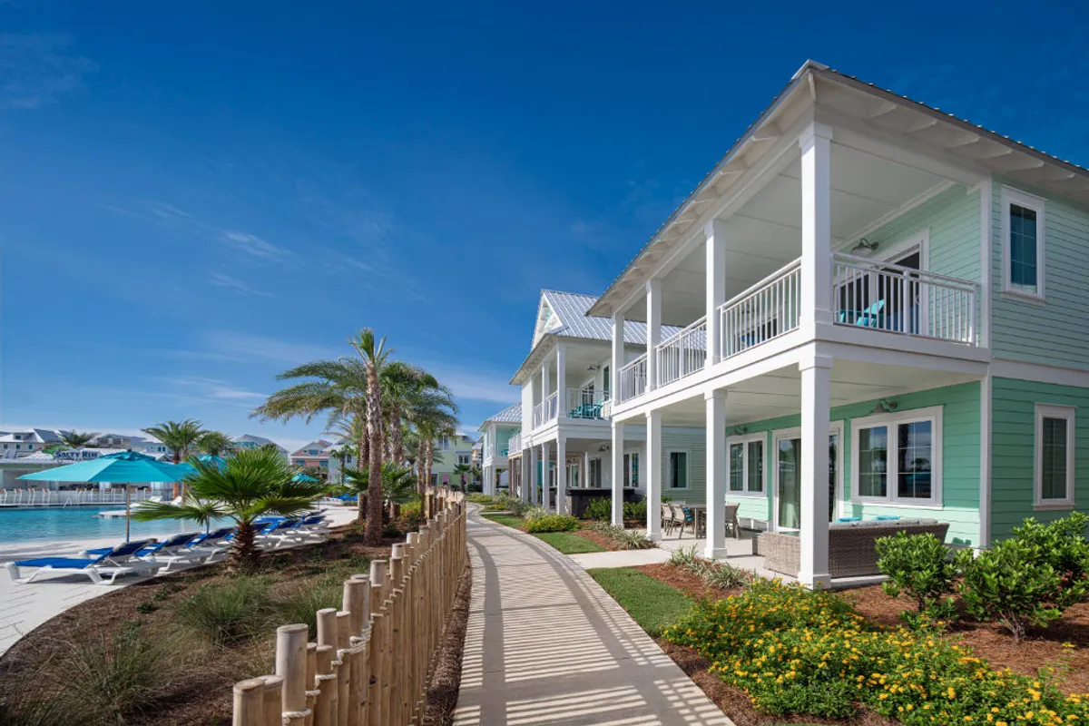 This image features a turquoise beach cottage with a spacious porch and balcony, set beside a resort pool with blue umbrellas and lounge chairs. The paved walkway is bordered by tropical plants and palm trees, with similar cottages visible in the background. The scene conveys the harmonious balance of private home comfort within a vibrant resort setting.