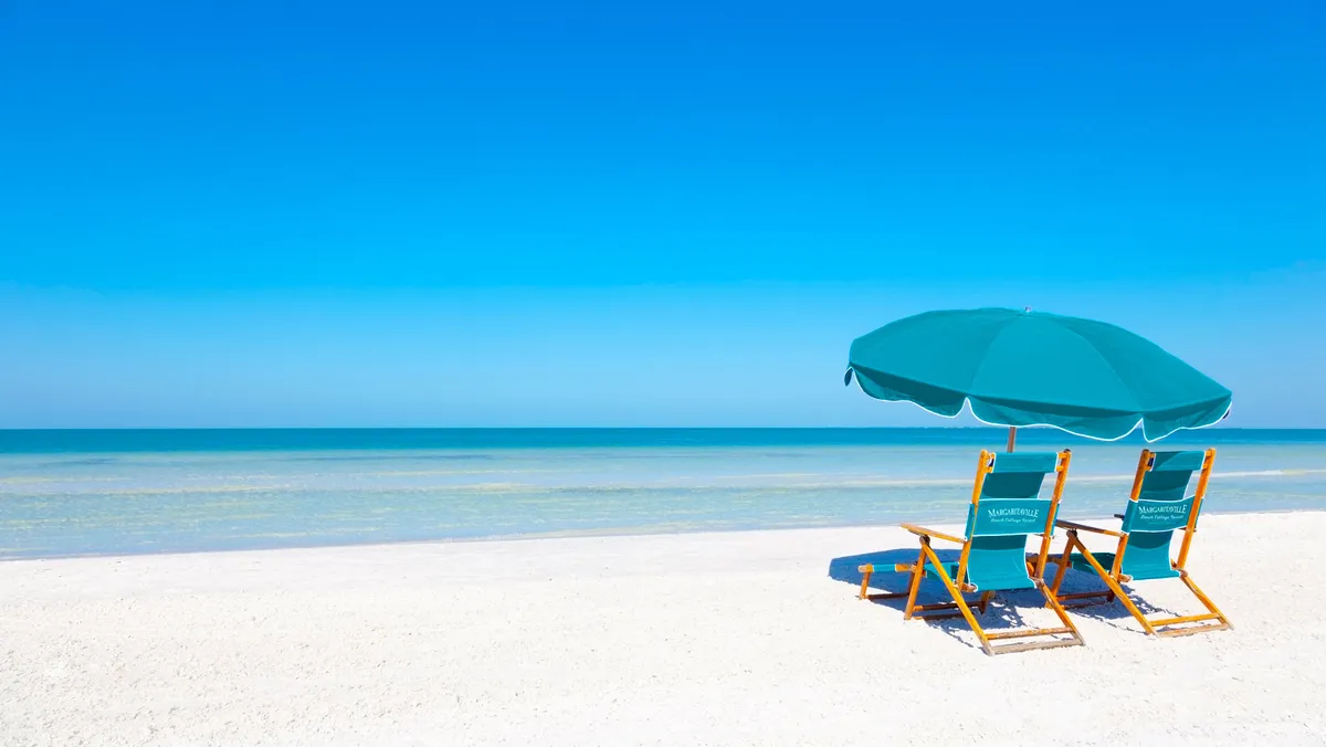 A pair of turquoise 'Margaritaville' beach chairs and matching umbrella are set on a pristine stretch of white sand, facing calm blue ocean waters under a clear sky. The setup is inviting for sunbathing, relaxing, or simply soaking in the serene beachfront environment.