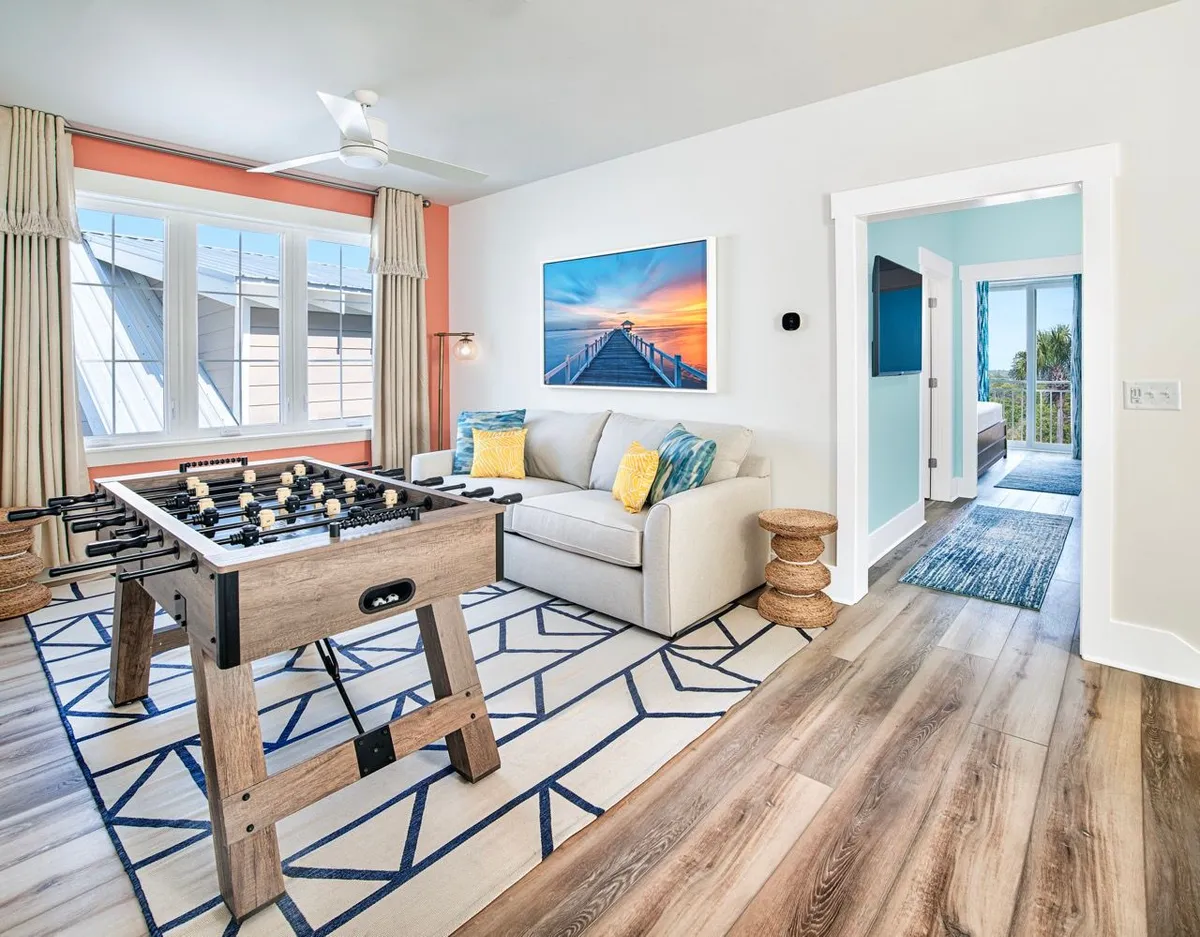 A well-lit, inviting game room with a foosball table set in the foreground, a cozy beige sofa with colorful cushions, large window letting in abundant natural light, and vibrant wall art depicting a sunset pier. The overall mood is relaxed and playful, ideal for family gatherings or group entertainment.