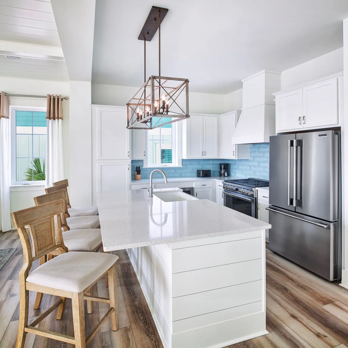 The focal point is a bright, modern kitchen with a large white island featuring a built-in sink, complemented by a stainless steel refrigerator and eye-catching blue subway tile backsplash. Wooden bar stools with upholstered seats line the island, suggesting a space for casual meals, conversations, or breakfast gatherings. The overall atmosphere is airy, fresh, and conducive to connection.