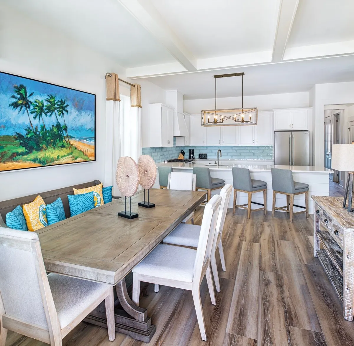 This image highlights a spacious, inviting dining area featuring a large wooden table adorned with decorative sculptures, set against the backdrop of a coastal-themed interior. The modern kitchen is visible with white cabinetry, a striking blue tile backsplash, and comfortable bar seating with grey upholstered stools. The scene is awash in natural light, reinforcing the cheerful, welcoming vibe.