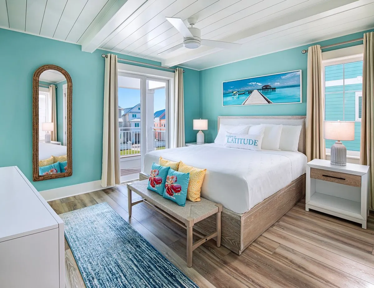 A spacious king bedroom featuring a plush bed with a 'Latitude' pillow, coastal artwork above the headboard, a large mirror with a woven frame, and balcony doors that open to a private outdoor view. Natural light floods the room, creating a fresh and tranquil atmosphere ideal for relaxation and rejuvenation.