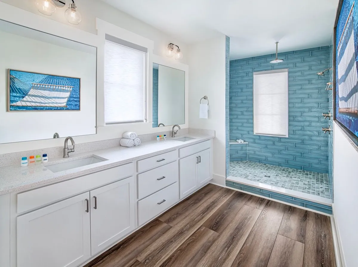 A bright, spa-inspired ensuite bathroom with a double vanity, large mirrors, white cabinetry, and a walk-in shower lined with blue subway tile and a rainfall showerhead. The wood-style flooring and coastal artwork accentuate the fresh, relaxing ambiance.