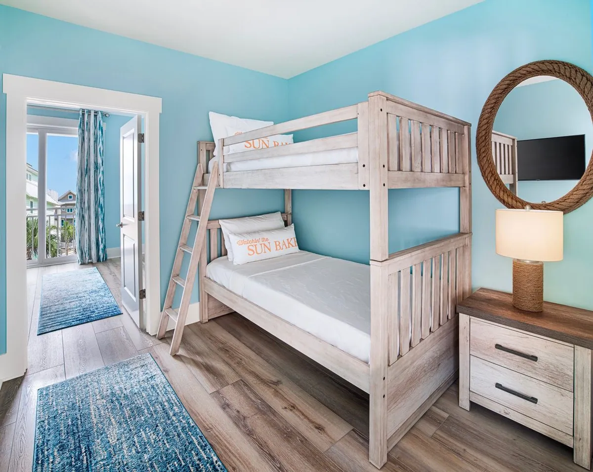 A cheerful bunk alcove with a wooden bunk bed featuring sun-themed pillows and blue walls, complemented by a round wicker mirror and a door that leads to a balcony with partial Gulf views. The room is bright and family-friendly, perfect for children or groups seeking a fun and restful space.