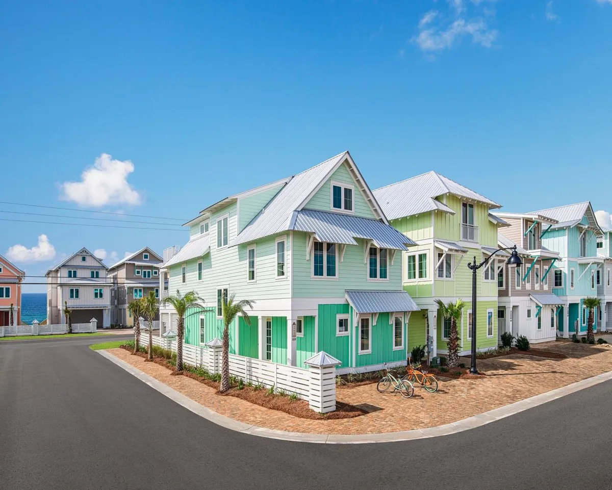 Rows of cheerful pastel beach cottages with metal roofs line a palm-shaded street, offering glimpses of the ocean in the background. Bicycles and a paved sidewalk invite exploration, creating a lively neighborhood feel that hints at easy access to both the beach and resort amenities.